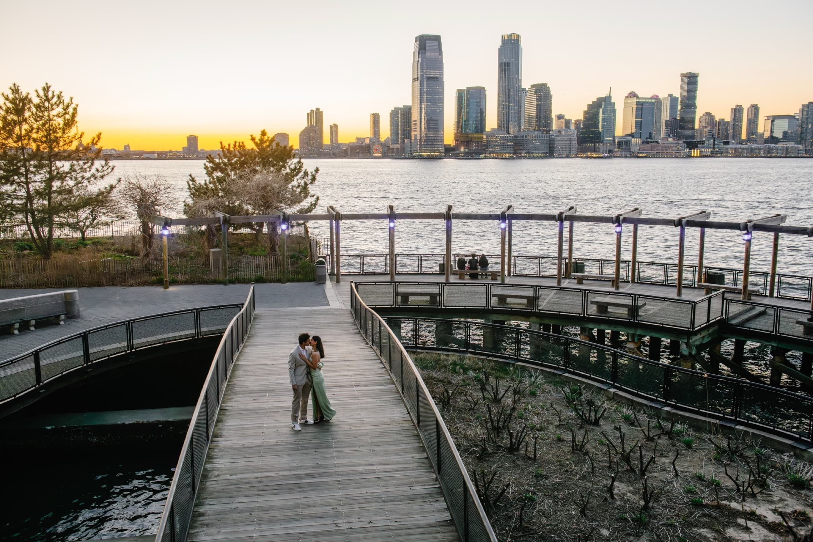 Krati and Shaan kissing on a waterfront boardwalk at sunset